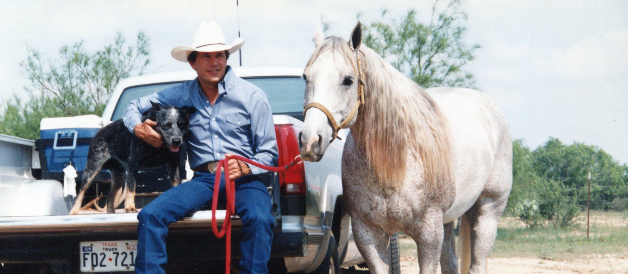 George Strait sits on the tailgate of the pickup petting a blue heeler dog and holding the lead rope of a grey horse standing beside the truck.